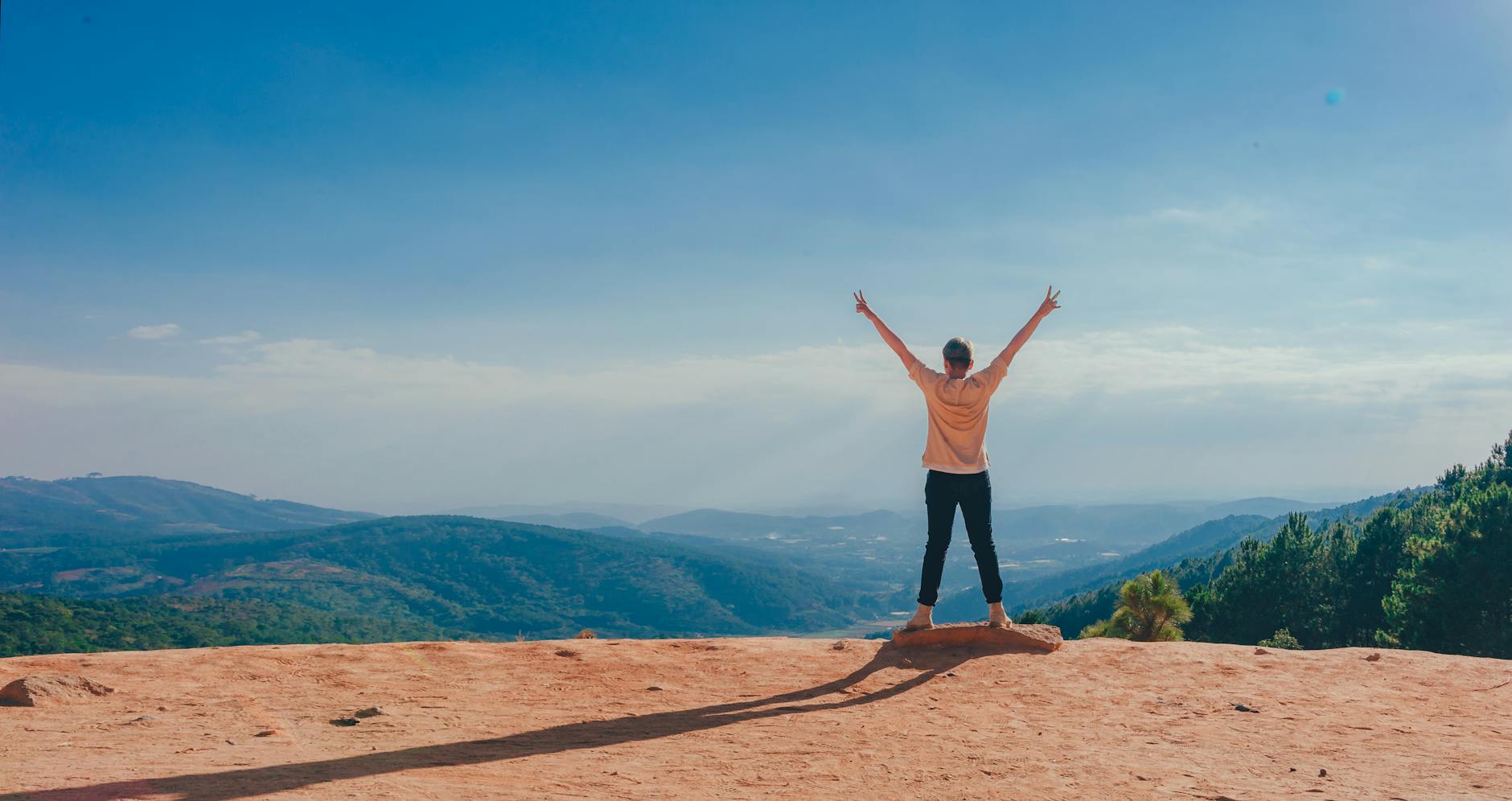 Image of an individual standing at the edge of a cliff facing a verdant canyon, back to the camera with arms outstretched and each hand holding up fingers in a V for victory sign.

Photo by Min An on Pexels.com
